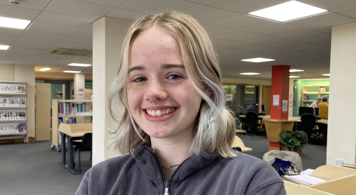 Young woman with shoulder length blonde hair, smiling to camera,  wearing grey fleece top and pictured in library with book shelves and desks in background.